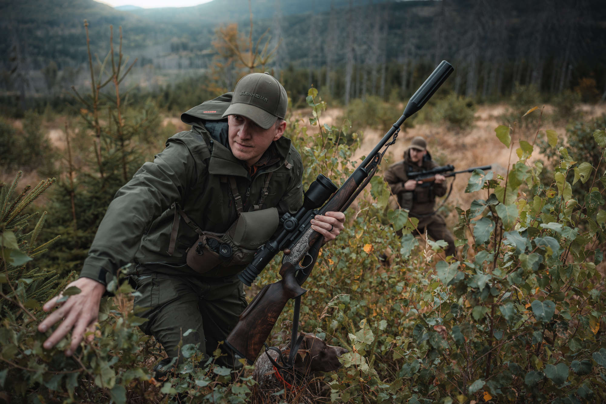 Hunter moving through scrub with a Hikmicro Stellar 4.0 scoped rifle, second hunter in the background
